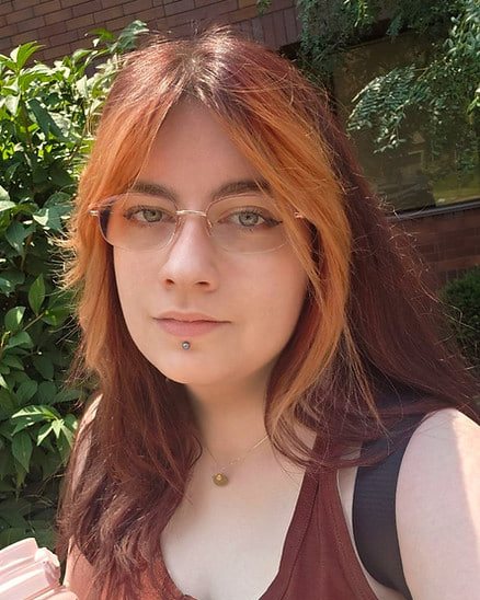 Destiny outside on a sunny day, in glasses and a rust-coloured top, her red hair down.