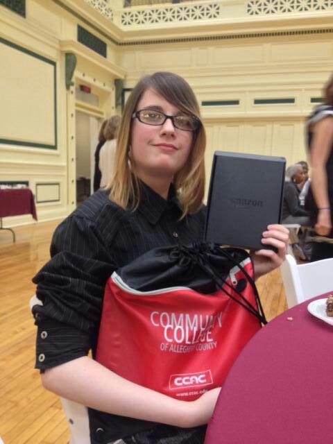 Destiny smiling in glasses, holding up her award and a red Community College of Allegheny County bag.
