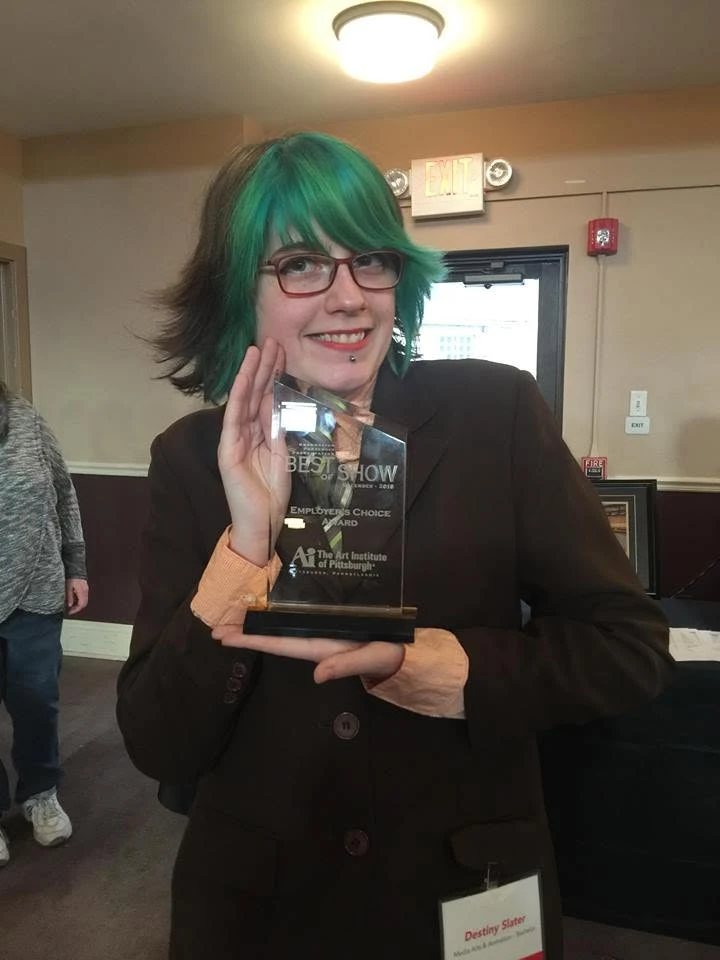 Destiny with green hair and glasses, smiling and holding her glass Best of Show award.