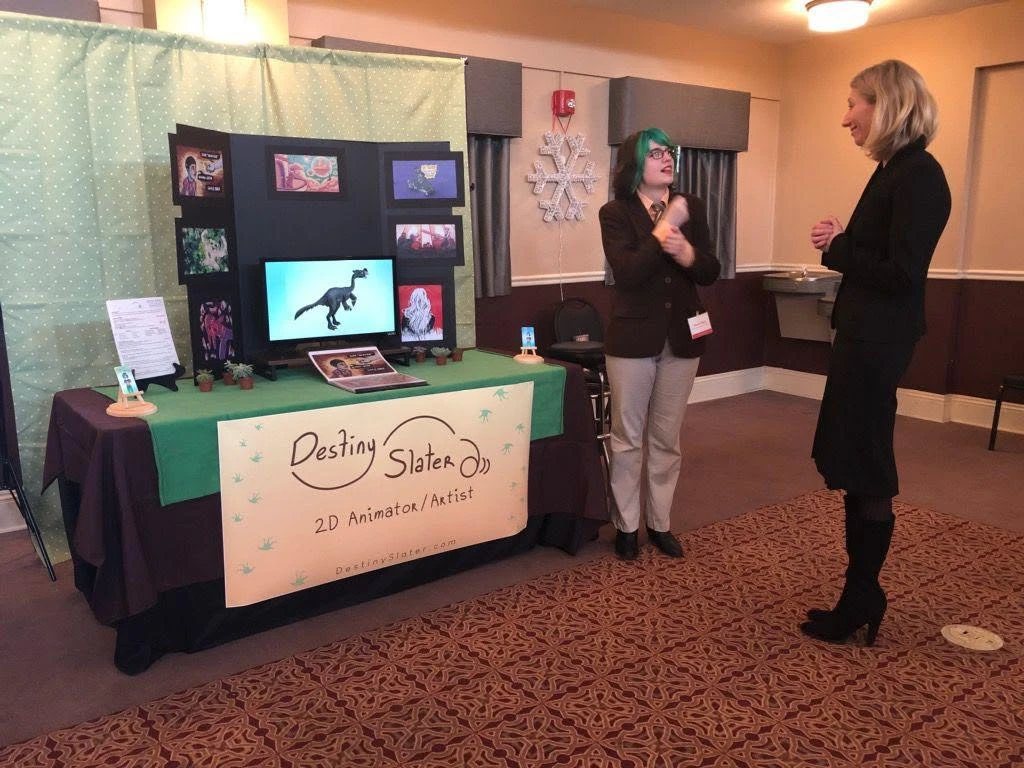 Her portfolio show table, with her art on screens and a banner reading Destiny Slater, 2D Animator, while she signs with a visitor.