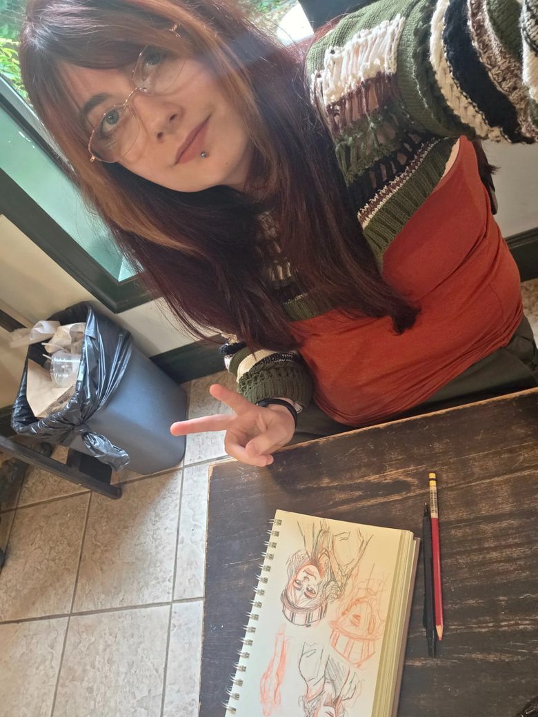 Destiny in glasses and a striped sweater, smiling and making a peace sign over her open sketchbook at a café table.