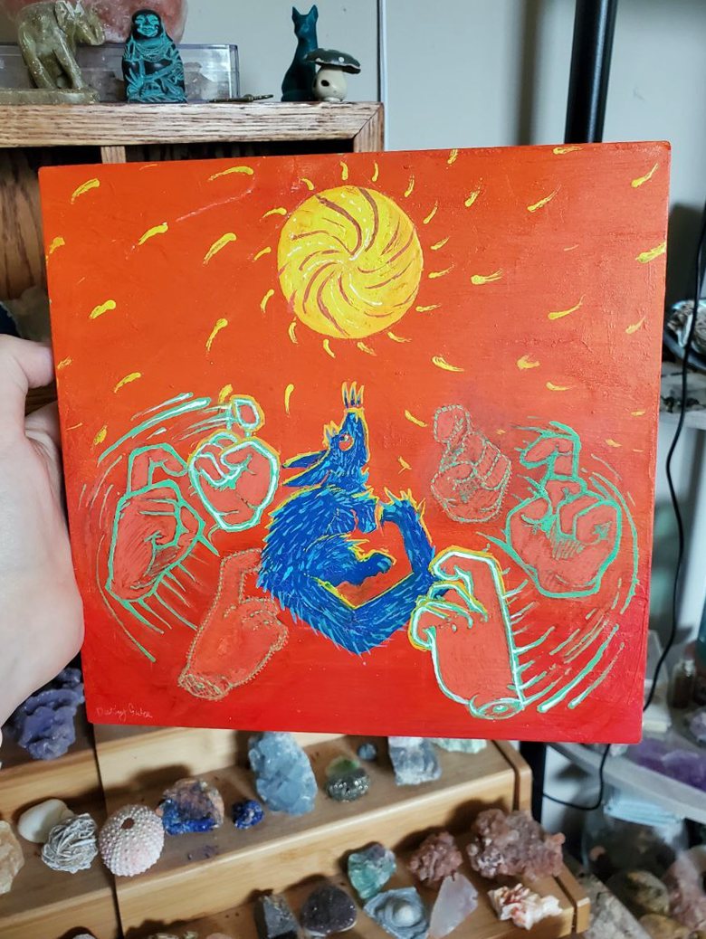 A hand holds up the finished square painting indoors, in front of shelves of crystals and small figures. The painting is red-orange, with a swirling yellow sun at the top, a furry blue creature in the center looking up at it, and hands outlined in glowing green around it signing CHANGE.
