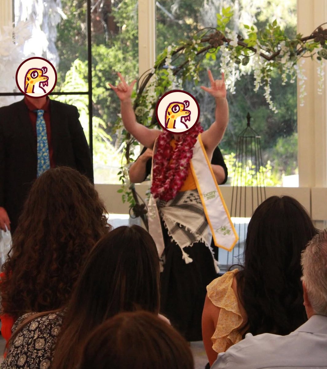 A graduate in a flower lei and a white stole stands at the front of a room and raises both hands in the ASL "I love you" sign. The backs of the audience's heads are in the foreground.