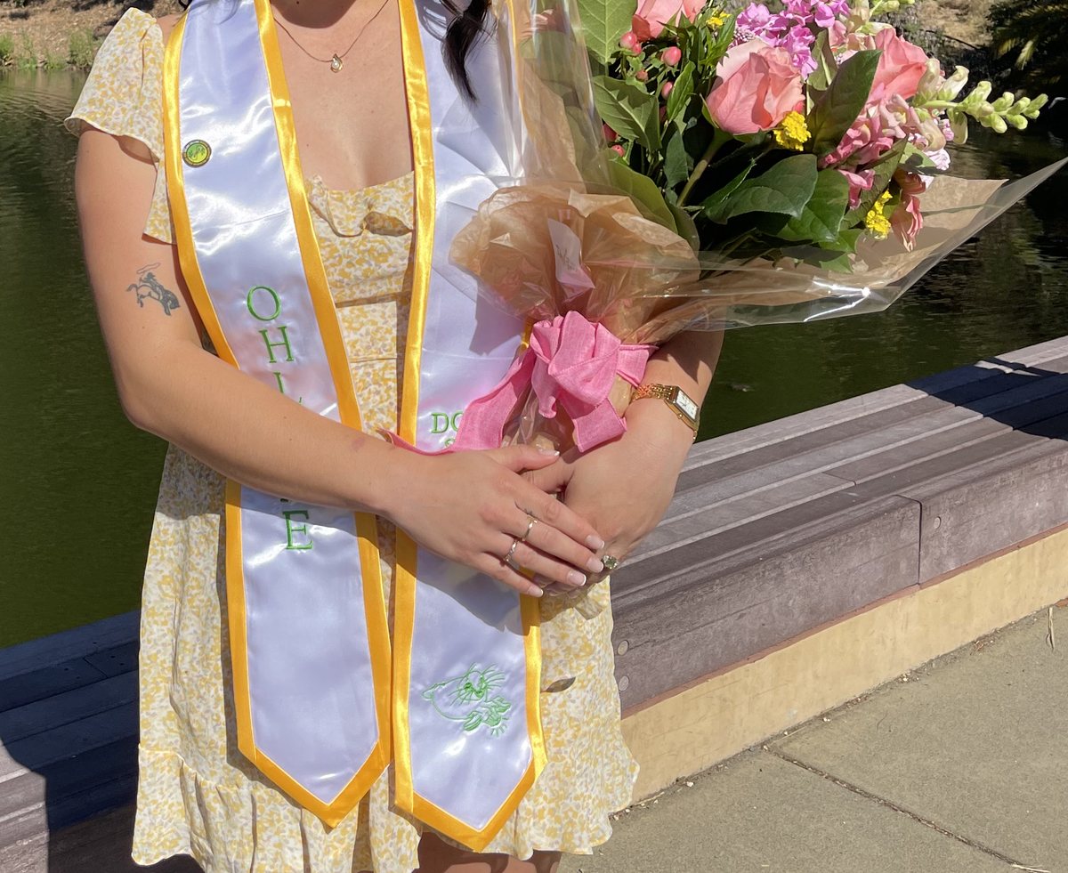 A graduate stands outdoors beside a pond, holding a bouquet of pink roses. The graduate wears a white stole with gold edges over a light dress.