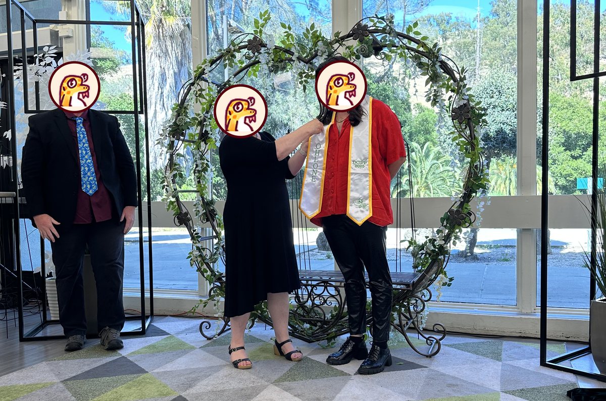 A graduate in a red top stands in front of the round flower arch while another person places a white stole with gold edges on them. A man in a suit stands to the left.