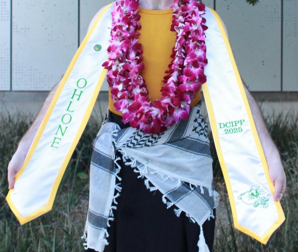 A graduate holds out both ends of a white stole with gold edges. One end reads "Ohlone" in green. The other end reads "DCIPP 2025", and below it Sally the Seal is embroidered in green thread, signing SEAL.