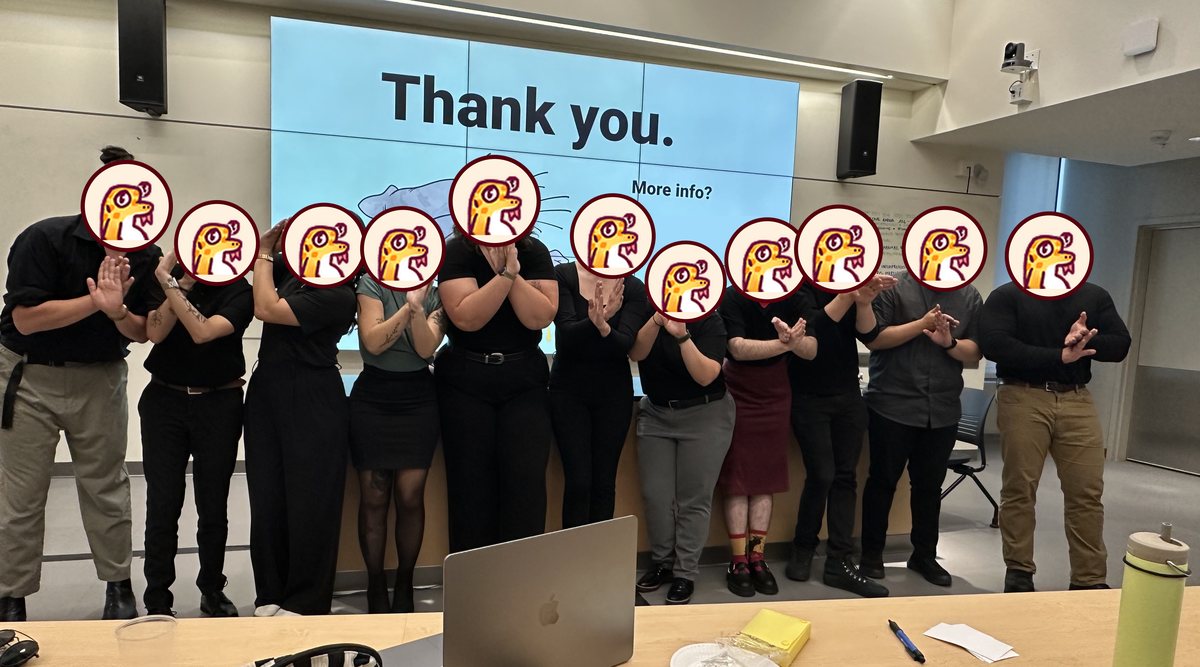 Eleven students dressed in black stand in a row in front of a big screen that reads "Thank you.", with Sally's drawing on the slide. Each student holds both hands in front of their chest, signing SEAL.