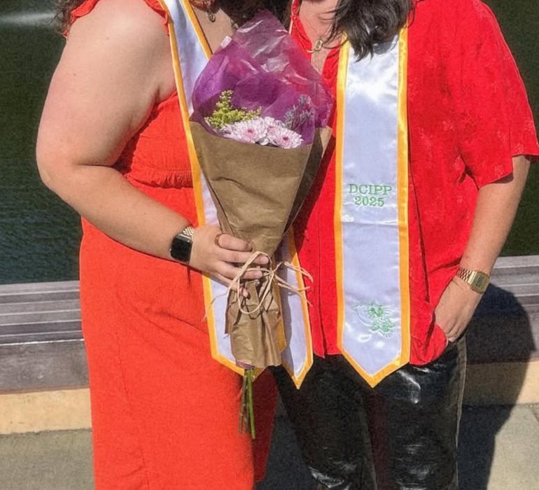 Two graduates stand outdoors in front of a pond, one in an orange dress and one in a red shirt. Both wear white stoles with gold edges, and one holds a bouquet of flowers.